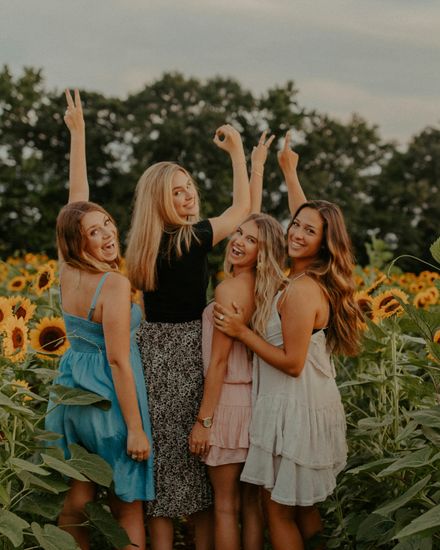 Vier junge Frauen stehen lachend mit erhobenen Armen in einem Sonnenblumenfeld