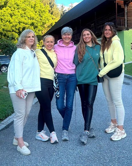 Fünf Frauen stehen Arm in Arm und lachend vor einem alten Holzhaus in der Abendsonne